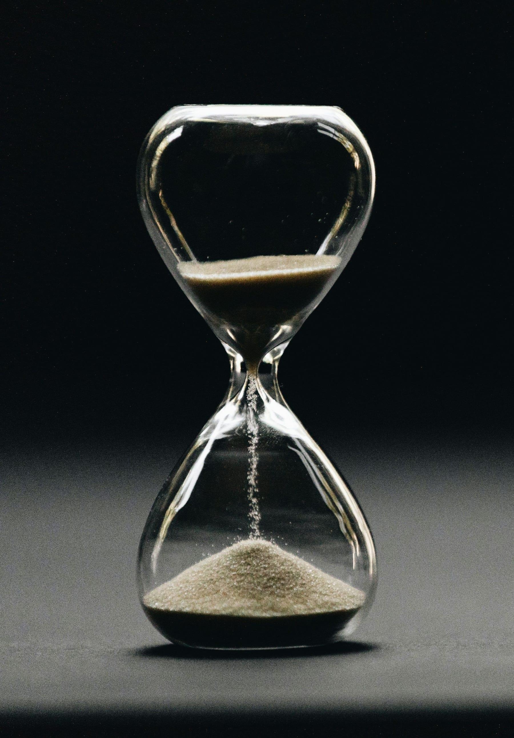 A glass hourglass with fine sand running through it, softly lit against a dark background