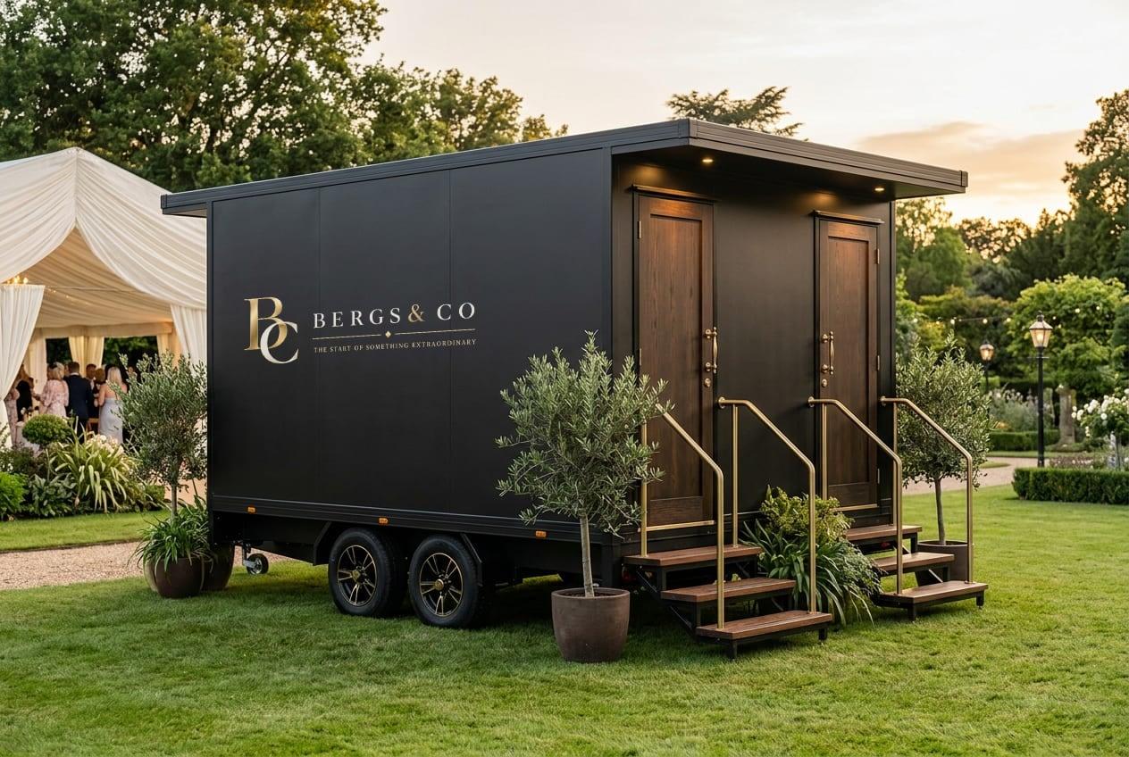 Black luxury mobile toilet trailer at a garden event, its side panel carrying the gold Bergs & Co lockup, with timber doors, brass handrails and olive trees